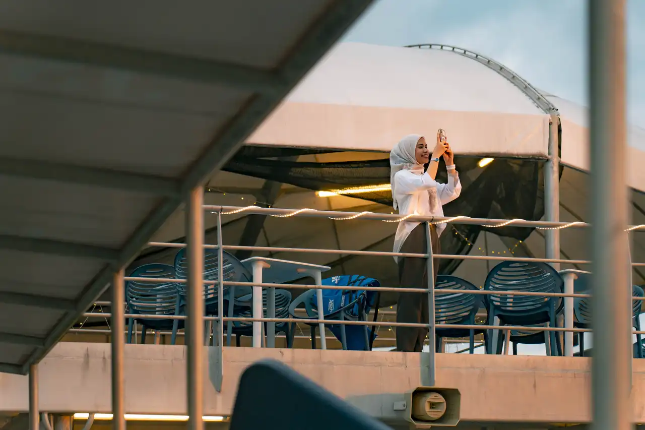 Woman in hijab taking a photo with her phone on an outdoor deck with string lights and stacked chairs.