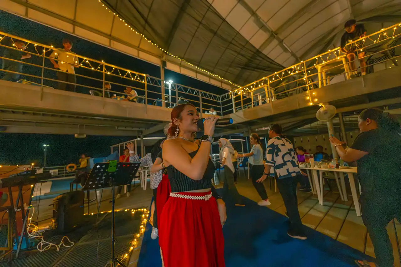 Woman singing into a microphone at a lively outdoor evening event with guests and string lights.