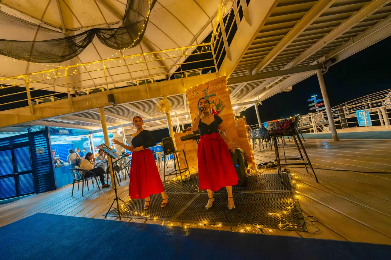 Two singers performing on a deck at night with festive lights and a seated audience in the background.