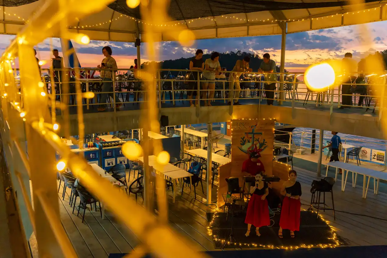 Outdoor multi-level dining area with string lights and live music performance at sunset.