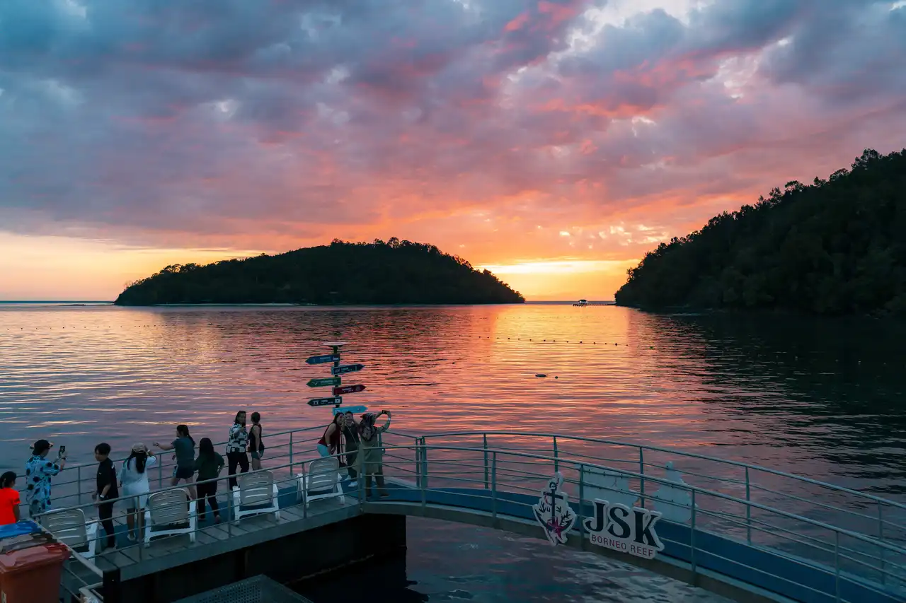 Sunset over a tranquil sea with people on a pier, featuring the JSK Floating Island sign.